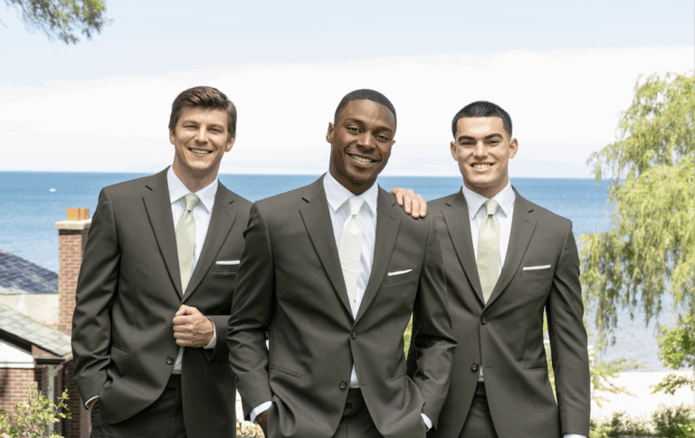 Three men in matching dark suits with light green ties stand outdoors, smiling, with a scenic view of trees, rooftops, and a large body of water in the background. One man has his hand on another’s shoulder. Should I rent or buy my suit or tuxedo?